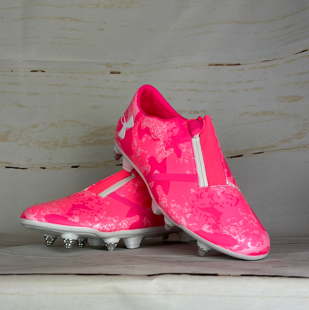 A pair of pink camouflage pattern Under Armour football boots with studs, displayed against a wooden background.