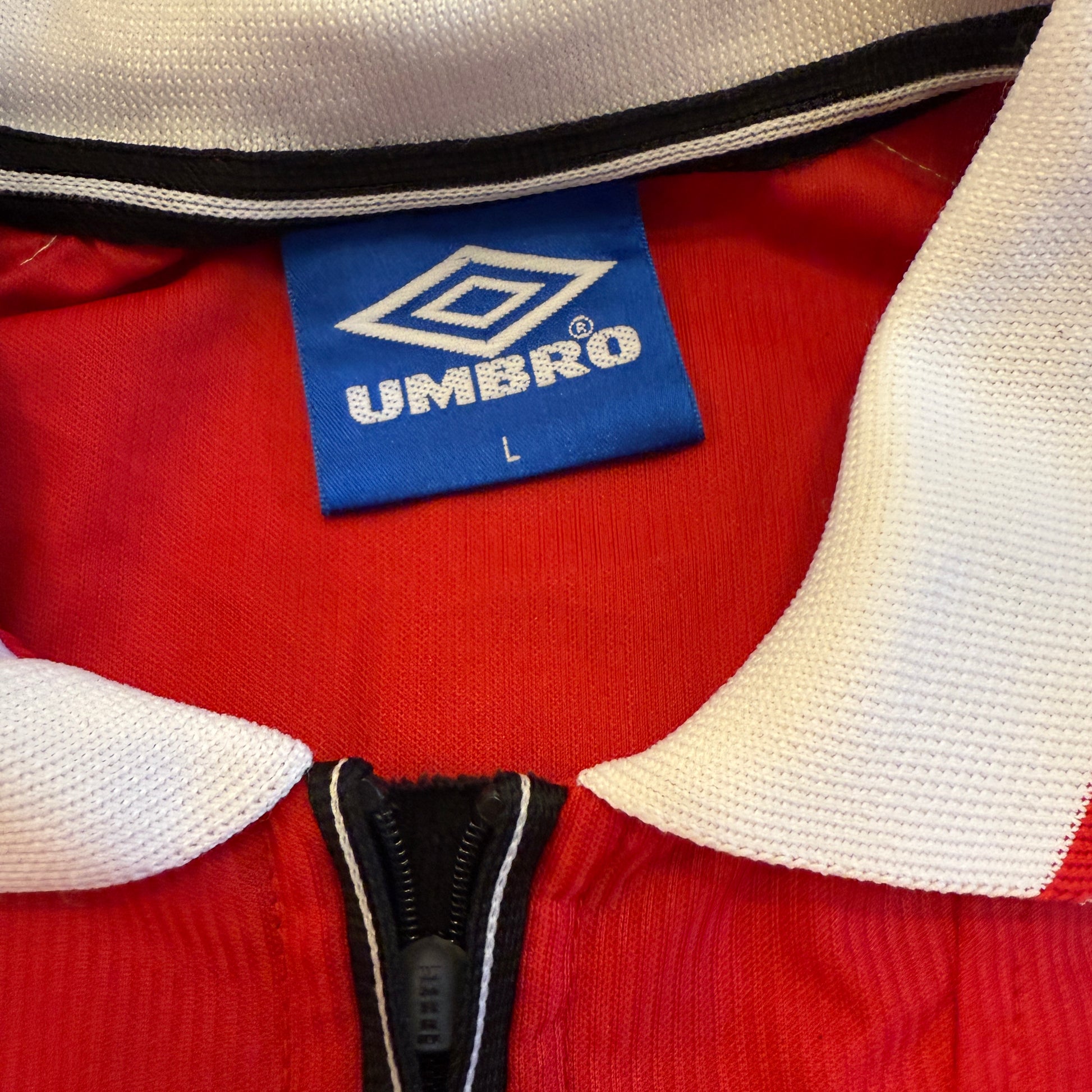 Close-up of a red jacket with a white collar and black zipper, featuring an Umbro label.