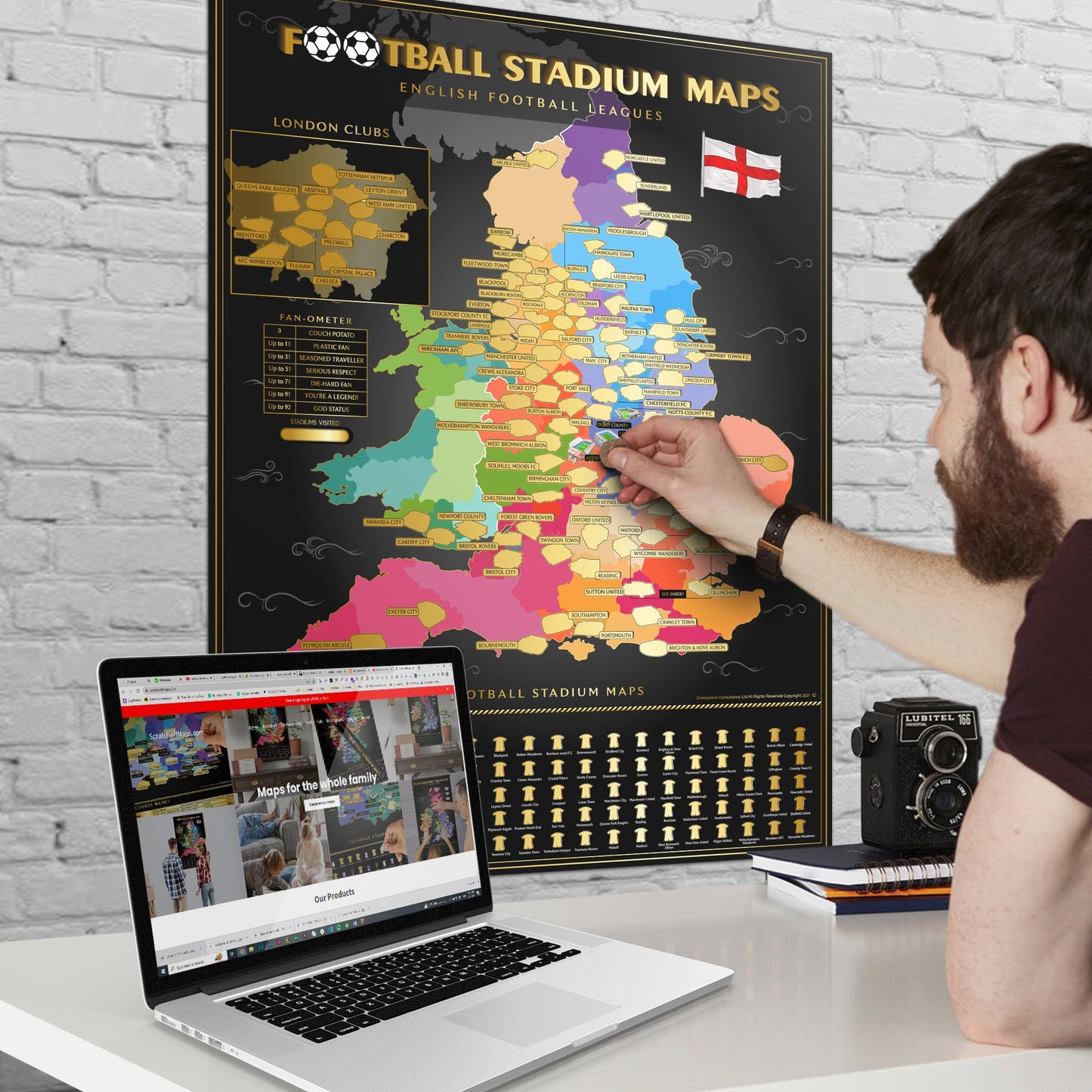 A person scratching off a map of football stadiums in England on a wall, with a laptop open on a desk.