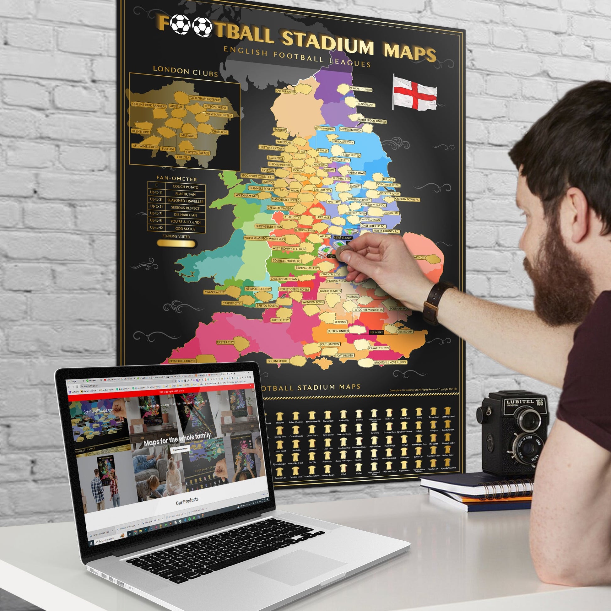 A person scratching off a map of football stadiums in England on a wall, with a laptop open on a desk.